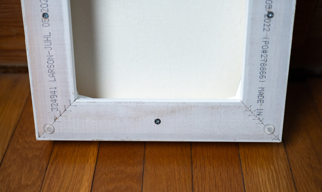 Back of a white-framed canvas artwork, showing staples, screws, and manufacturer markings, on wooden floor.