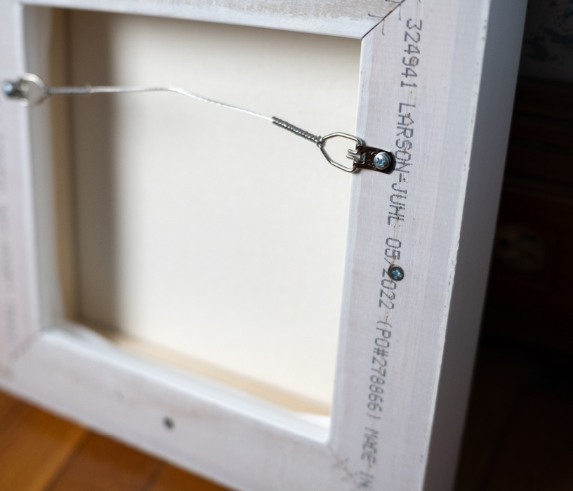 Back of a white-framed canvas artwork, showing staples, screws, and manufacturer markings, on wooden floor.