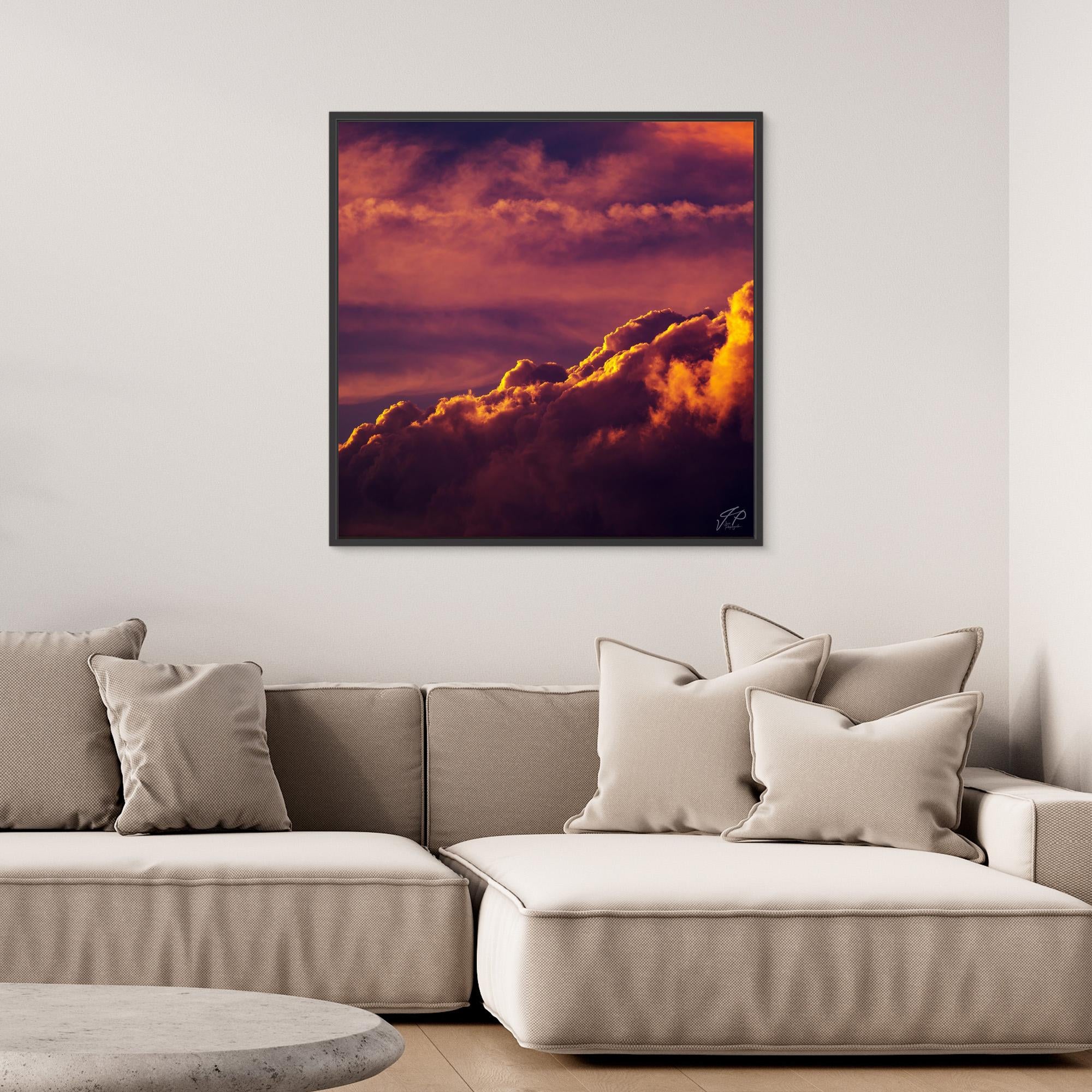 A framed photograph of dramatic, glowing sunset clouds hangs above a beige sectional sofa with pillows.
