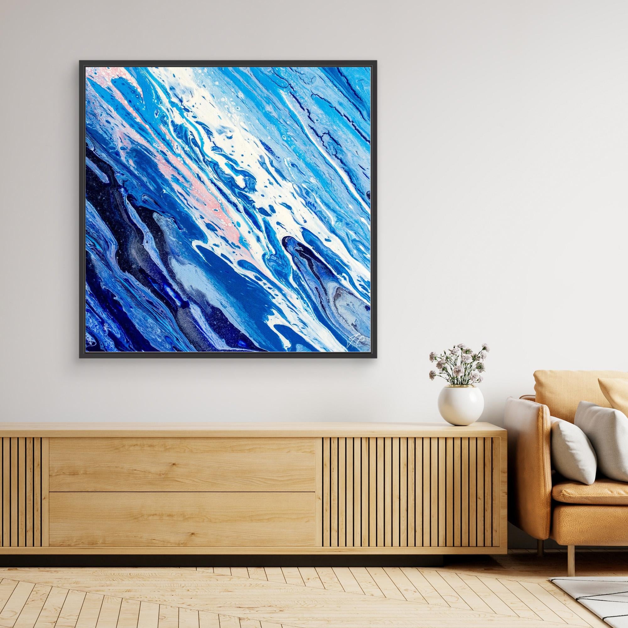 Blue and white abstract painting above wooden console with beige armchair and white vase of flowers.